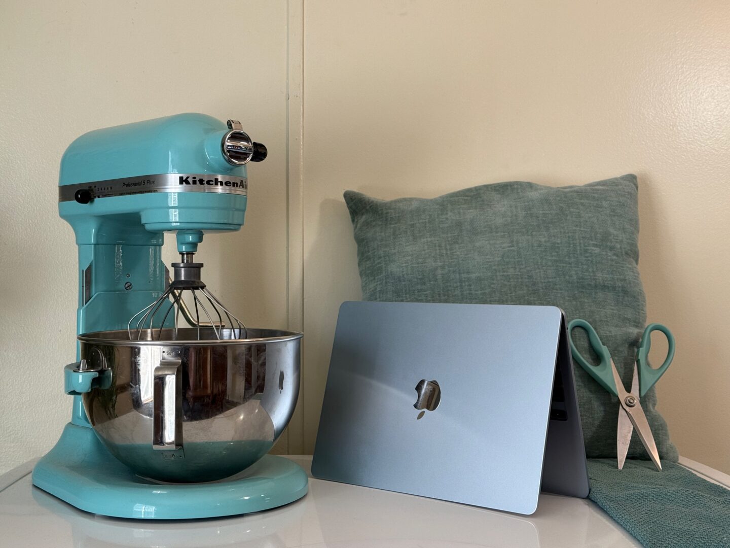MacBook Air M4 – Blue Comparison Sky blue M4 MacBook Air next to an aqua sky KitchenAid stand mixer, light blue pillow, scissors and towel