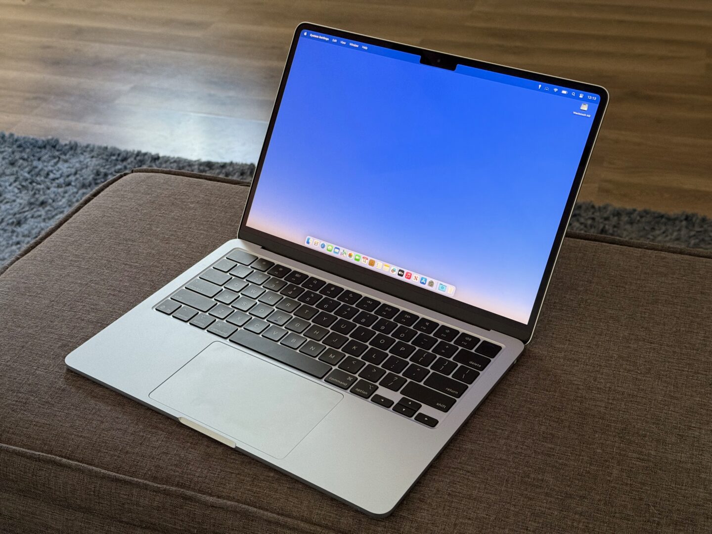 MacBook Air M4 – Ottoman Blue MacBook Air sitting on a gray linen ottoman on a blue fuzzy rug on a gray laminate floor